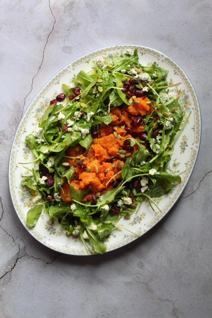 Orange butternut squash sits upon a bed of arugula with pumpkin seeds, blue cheese, and craisins.