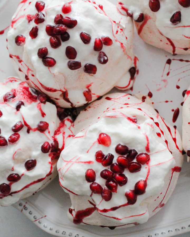 Pomegranate mini pavlovas on a white tray covered in whipped cream and pomegranate syrup and seeds.