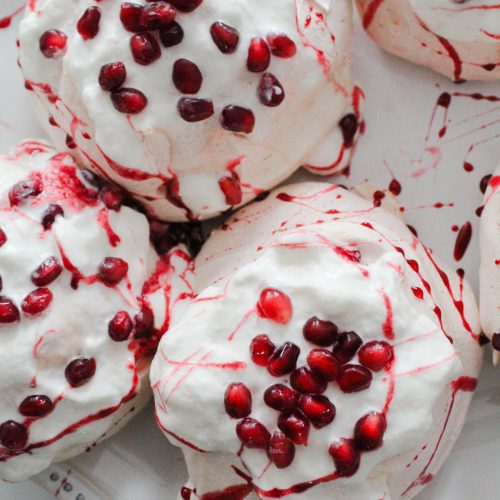 Pomegranate mini pavlovas on a white tray covered in whipped cream and pomegranate syrup and seeds.