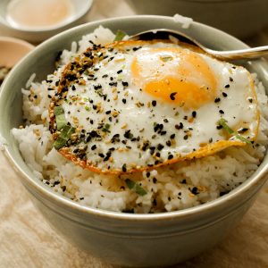 A green bowl sitting upon a biege cloth filled with rice covered in sesame sauce, a fried egg, and furikake.