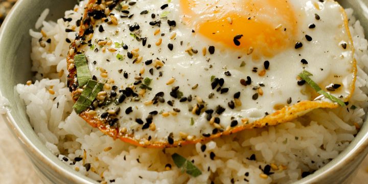 A green bowl sitting upon a biege cloth filled with rice covered in sesame sauce, a fried egg, and furikake.
