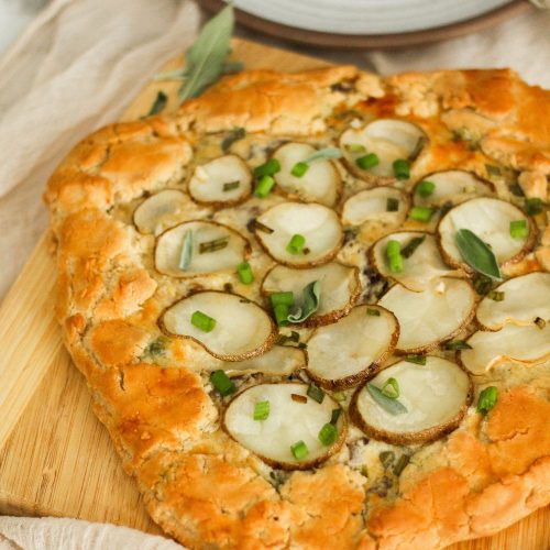 Golden savory galette covered in potatoes and green onions sitting on a wooden cutting board.