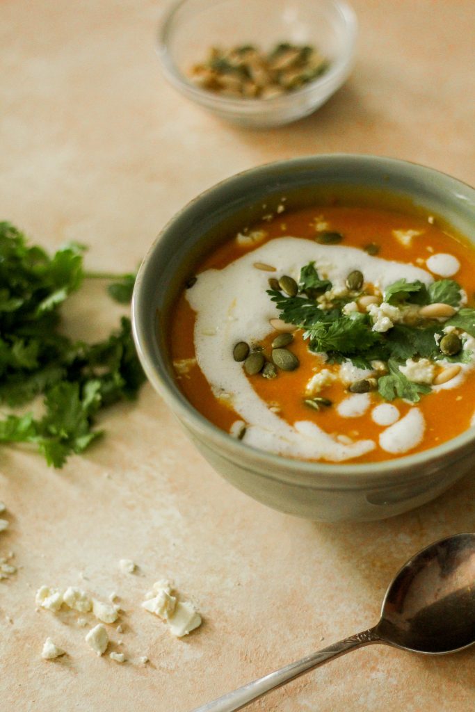 A green bowl filled with butternut squash bisque topped with cilantro, pine nuts, pumpkin seeds, and feta.